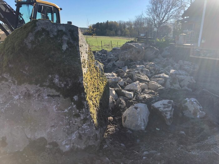 Stor sten mossig framför, stenröse bakom, traktor till vänster, soligt landskap, träd, byggmaterial i bakgrunden.