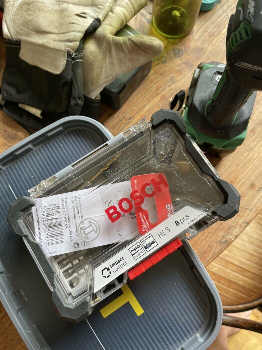 Bosch borrbitssats i förpackning, batteridriven borrskruvdragare och arbetshandskar på träyta.