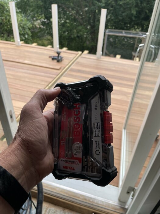 En hand håller en Bosch borrbitsförpackning med terrassbygge i bakgrunden.