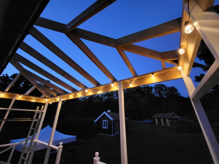 Träpergola med ljusslinga i skymningen, utsikt över trädgård och litet hus.