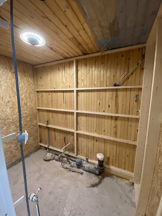 Under renovering, rörmokeri synligt, träpanel, OSB-skivor, betonggolv, elektrisk utrustning, oavslutat rum, takbelysning.