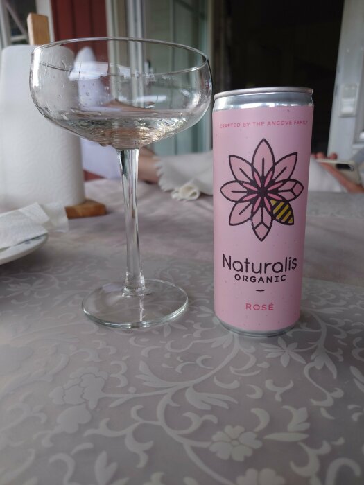 Ett vinglas med rester av vätska och en rosa burk märkt "Naturalis Organic Rosé" på ett bordsduksmönstrat bord.