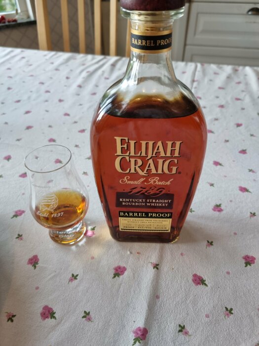 Elijah Craig bourbonflaska på blommigt duk, glas med whisky bredvid.