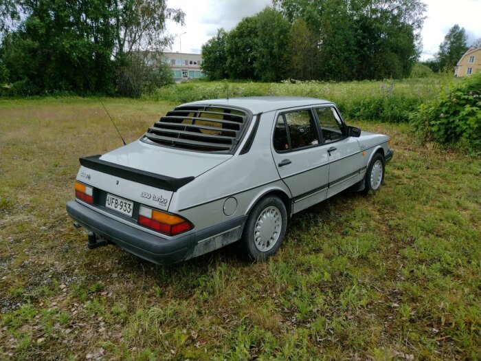 Klassisk silver Saab 900 Turbo parkerad på en övergiven gräsplätt, träd och hus i bakgrunden.