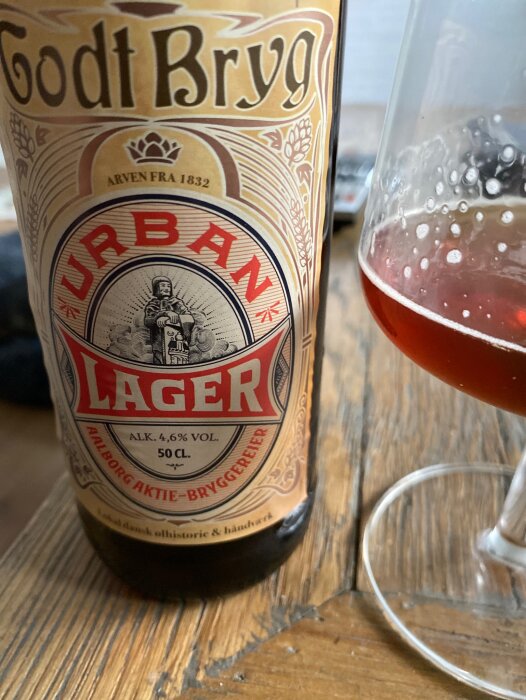 Ölflaska märkt "Godt Brygg" "Urban Lager", 4.6% vol, bredvid delvis fylld ölglas på träbord.