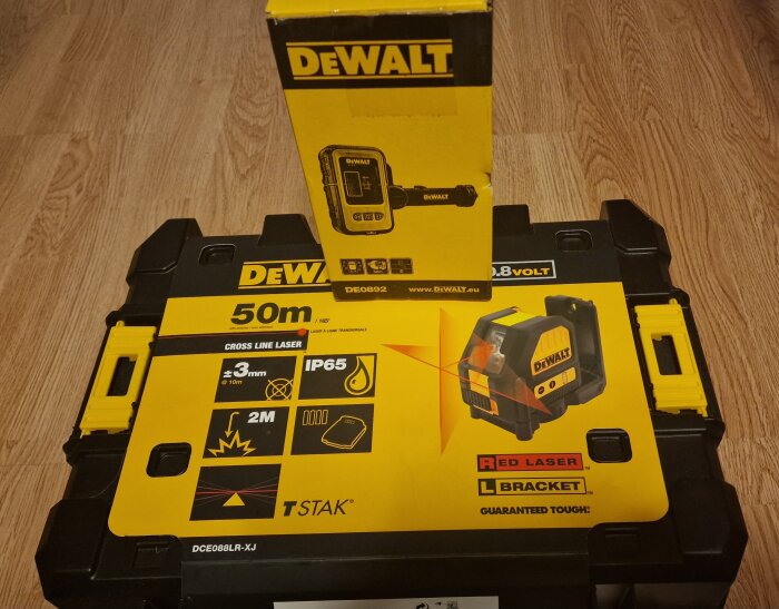 Dewalt korslinjelaser förpackning, gulsvart, 50m räckvidd, IP65, TSTAK-kompatibel, trälaminatgolv i bakgrunden.