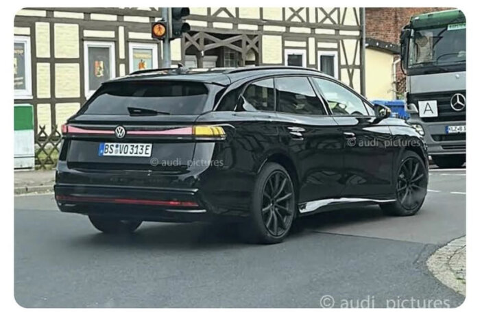 Ett svart elbil, antagligen Volkswagen, vid trafikljus bredvid företagskyltat lastfordon i urban miljö.
