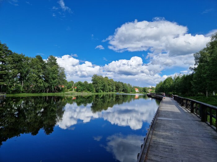 Trädbevuxen sjö med spegelreflektion, träbrygga, promenerande person, moln och klar blå himmel.