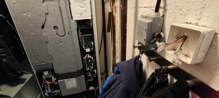 Öppnad elektronisk enhet, kablar, kontrollpanel, kläder, rörledningar och en opålitlig installation mot en vit vägg.