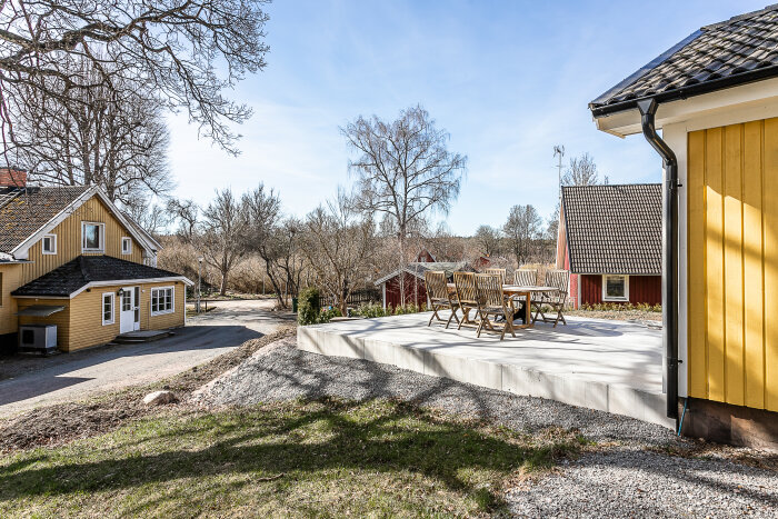 Trädgård med uteplats, trämöbler, gult hus, blå himmel, tidig vår, ingen snö, soligt, lugnt bostadsområde.