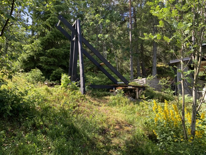 Svart träkonstruktion i skogsmark, växtlighet, soligt, gulblommor i förgrunden, del av byggnad synlig.