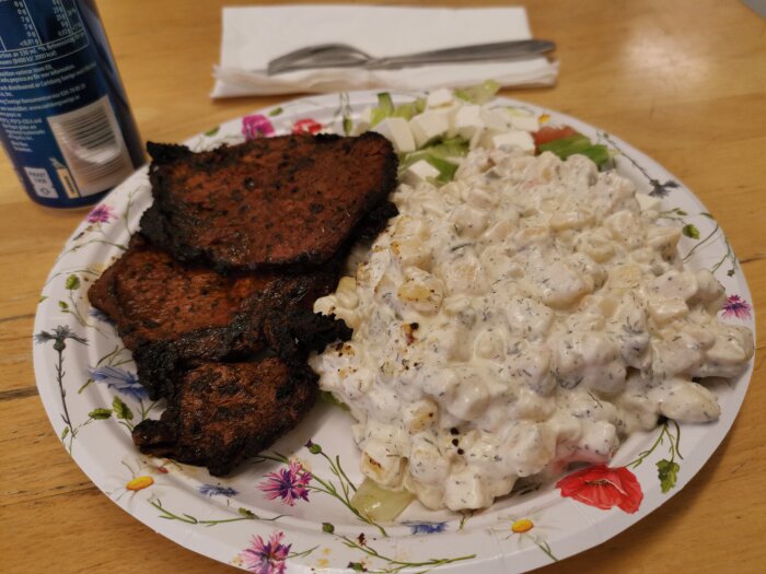 Grillade köttbitar, potatissallad, grönsallad på blommig tallrik med dryck och servett i bakgrunden.