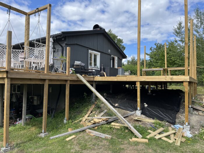 Ett svart hus med pågående altanbygge, trävirke, utemöbler och grönska i bakgrunden.