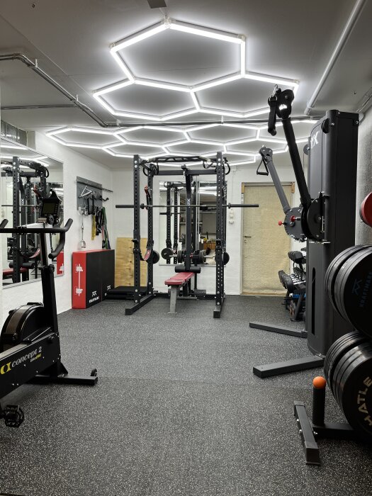 Modernt gymmed rum med utrustning för styrketräning, LED-takbelysning, roddmaskin och vikter.