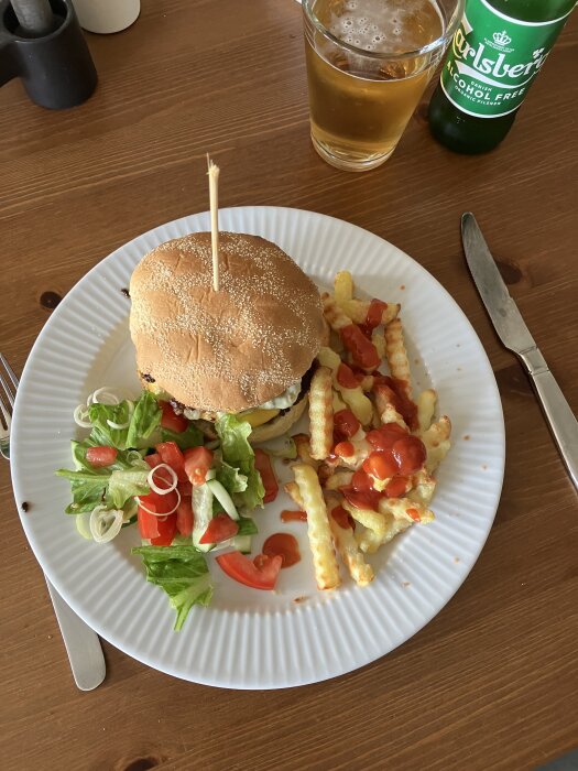 Hamburgare på tallrik med frasiga pommes och sallad, ölglas och alkoholfri ölburk bredvid.