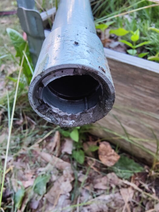 Slitet, blått metallrör med rost och smuts, närbild, utomhus mot otydlig grön bakgrund av gräs och löv.