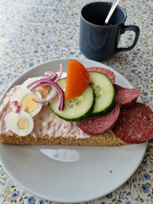 Ett smörgås med salami, ägg, gurka, rödlök och majonnäs på ett bord bredvid en kopp.