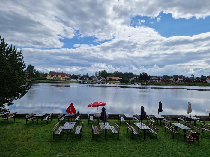 Molnig himmel över sjö med tomma stolar och bord, parasoller, trähus i bakgrunden.