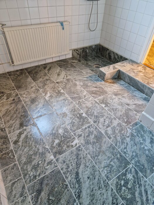 Ett badrum med grå marmormönstrade klinkers, duschhörna, vit radiator och vita väggkakel.