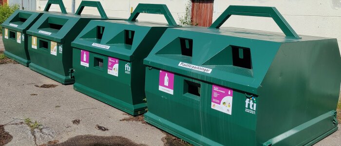 Gröna återvinningsstationer för förpackningar och tidningar utomhus, märkta för olika materialtyper.