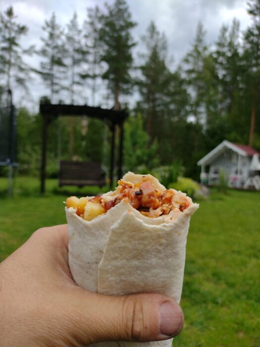 En hand håller en wrap med fyllning, natur och ett litet hus suddigt i bakgrunden.