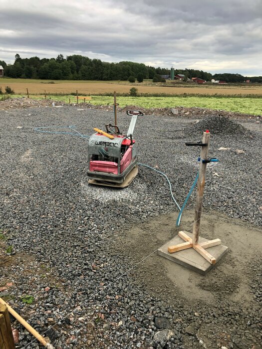 Markarbete med packningsmaskin, grus, stöd för nivellering och landsbygdsbakgrund med gård.