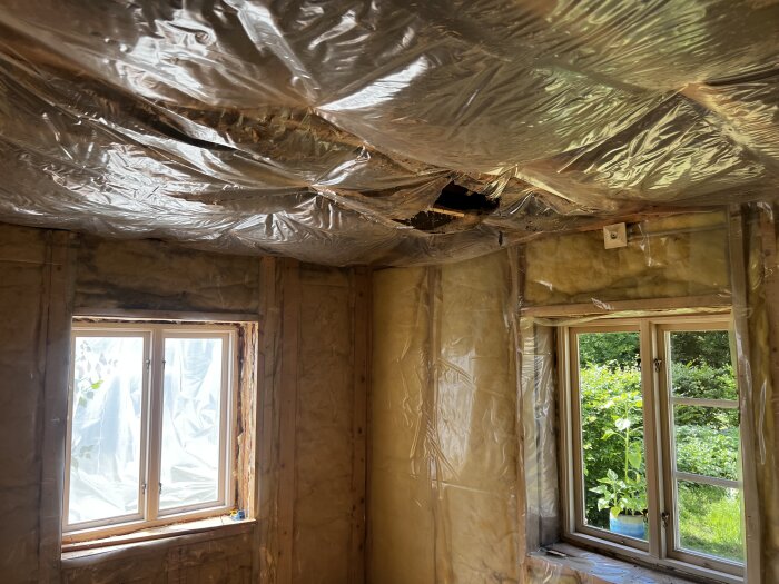 Renovering pågår, isolering synlig, plasttak hänger ner, skadat område i tak, oavslutat byggprojekt, två fönster mot grönska.