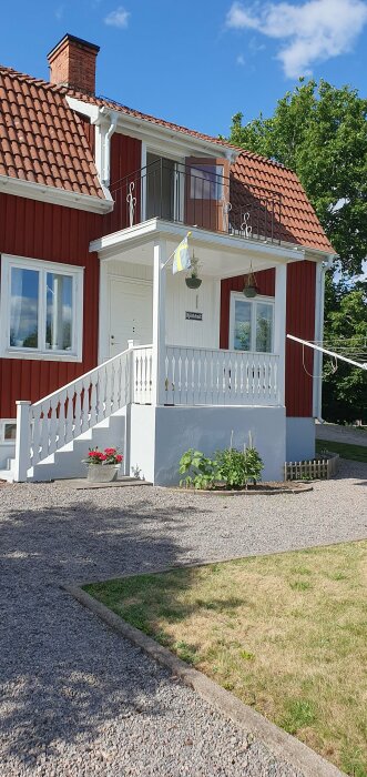 Rött trähus, vitt staket, balkong, svenska flaggan, gröna växter, blå himmel, sommardag, namnskylt "Björkhult".