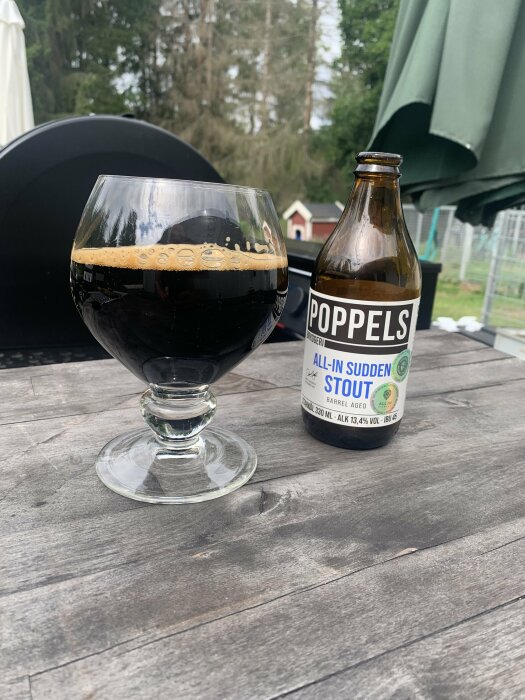 Ett glas med mörkt öl och en tom ölflaska märkt 'Poppels' på ett träbord utomhus.