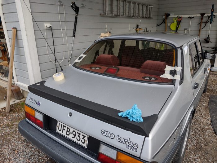 Silverfärgad Saab 900 Turbo, tejp på bakrutan, blå handskar, parkerad vid garage.