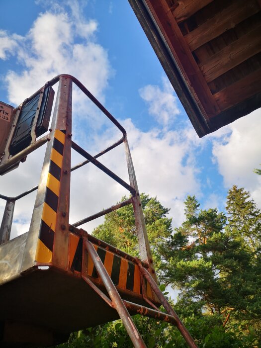 Stigande trappsteg mot blå himmel, vedbyggnad, industriell stege, gröna träd, moln.