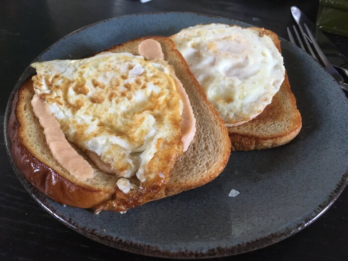 Två stekta ägg på rostat bröd med en korvskiva, serverat på en mörk tallrik.