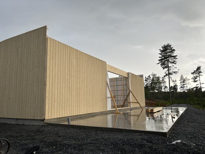 Modern träbyggnad under konstruktion, regnvåt grund, grus, skogsbakgrund med molnig himmel.