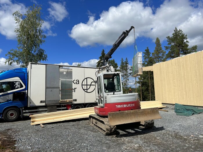 Minigrävare lastar byggmaterial nära lastbil vid byggarbetsplats, solig dag med blå himmel och träd.