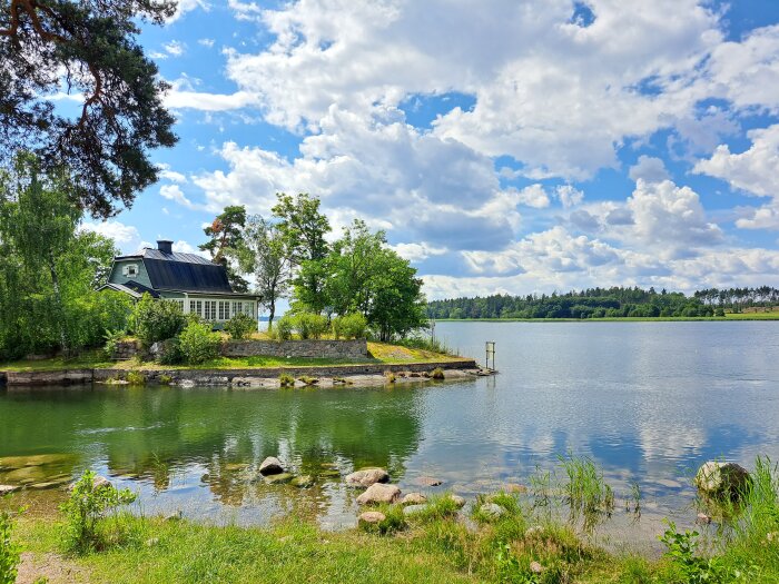 Idyllisk sjöutsikt, gröna träd, blå himmel med moln, traditionellt hus vid vattnet, avkopplande naturmiljö.