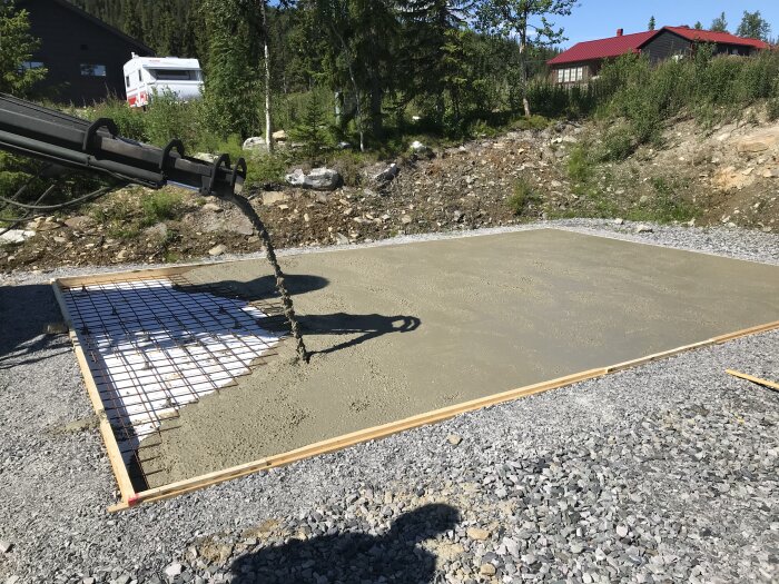 Betonggjutning i process, armeringsnät på marken, betongpumparm, soligt, hus och träd i bakgrunden.