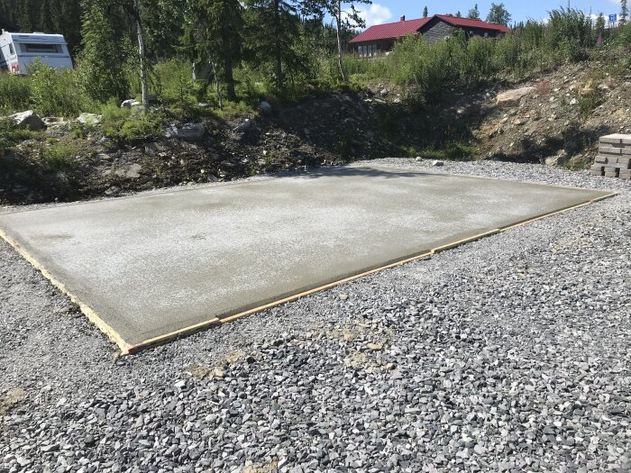 Nytillverkad betongplatta omgiven av grus, utomhus, dagtid, med skog och röd byggnad i bakgrunden.