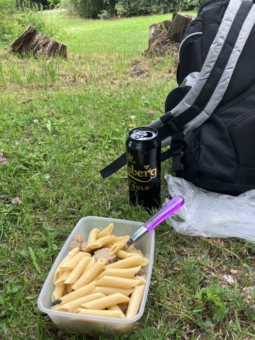 Utomhus picknick med pasta, energidryck och ryggsäck på en grön gräsmatta.