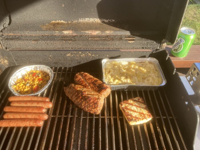 Grill med stekta köttstycken, korvar, majsblandning och gratäng. Burköl vid sidan. Utomhusmatlagning. Soligt väder.