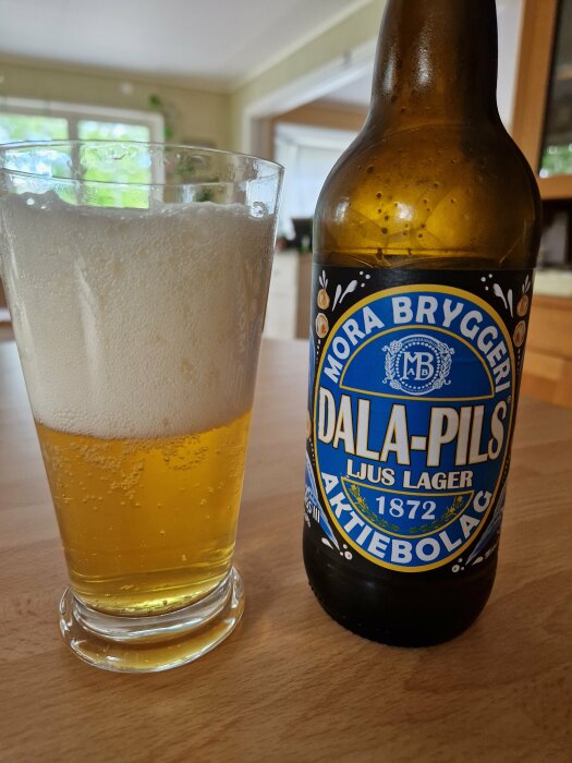 Ett glas med öl bredvid en flaska märkt "Mora Bryggeri Dala-Pils", inomhusmiljö.