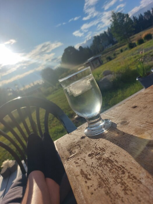 Ett glas på ett träbord, fötter i strumpor, solnedgång, avkopplande utomhusmiljö.