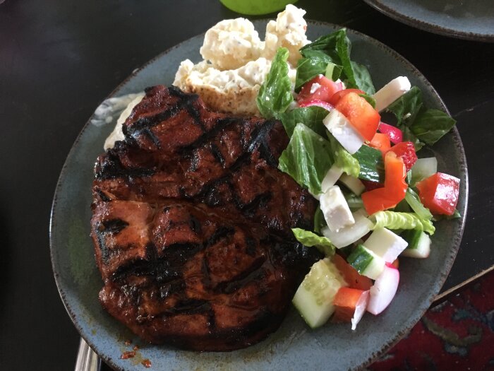 Grillad köttbit, sallad med tomat, gurka, lök, dressing, potatissallad på blått fat.