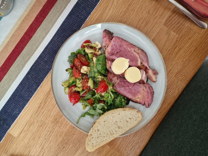 En tallrik med skivat kött, smör, sallad, tomat, avokado, brödbit, på träbord med färgglad löpare.