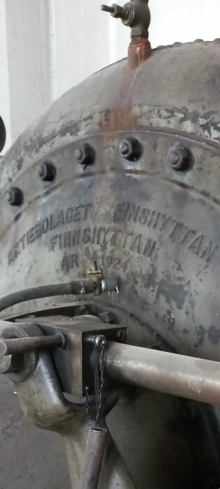 Gammal, rostig industriell maskindel med text "Nyttjebolaget Finshyttan år 1924", kopparfärgad ventil, skruvar, metalldetaljer.