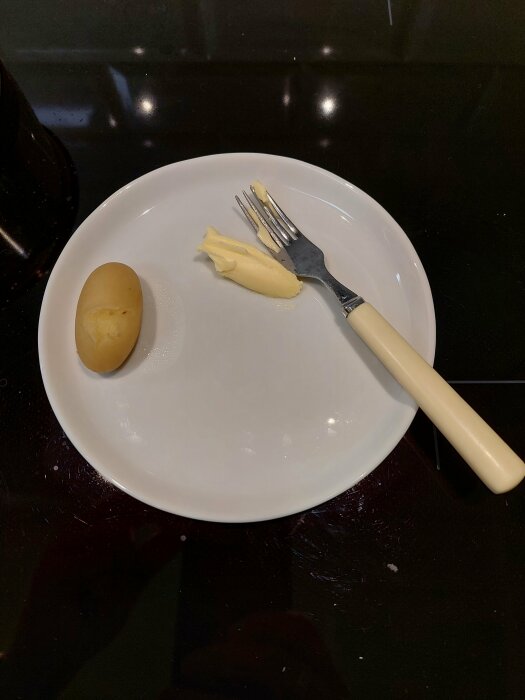 Vitt tallrik på svart yta med kokt potatis, bit smör och gaffel. Enkel, minimalistisk måltid.