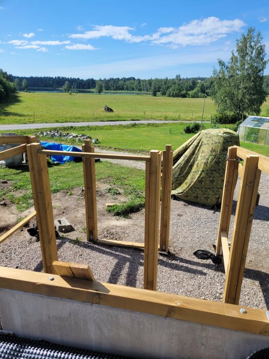 Trädgårdsprojekt, obehandlat trä, grönt landskap, molnig himmel, utomhus, oavslutad konstruktion, grusstig, tältduk, sommardag.