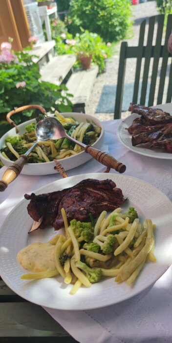 Uteplats med mat: grillade revbensspjäll, pasta, romanescobroccoli, sås, solljus, grönskande trädgård i bakgrunden.