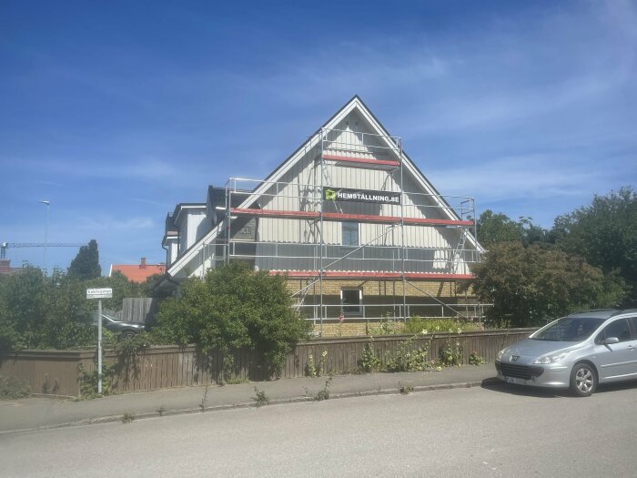 Modernt hus med triangulär form, ställningar, bil i förgrunden, blå himmel, träd och staket.