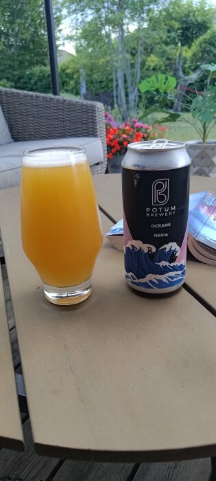 Ölglas med grumlig dryck bredvid en burk med etikett "OCEANS NEIPA". Utomhus med grönska och blommor i bakgrunden.