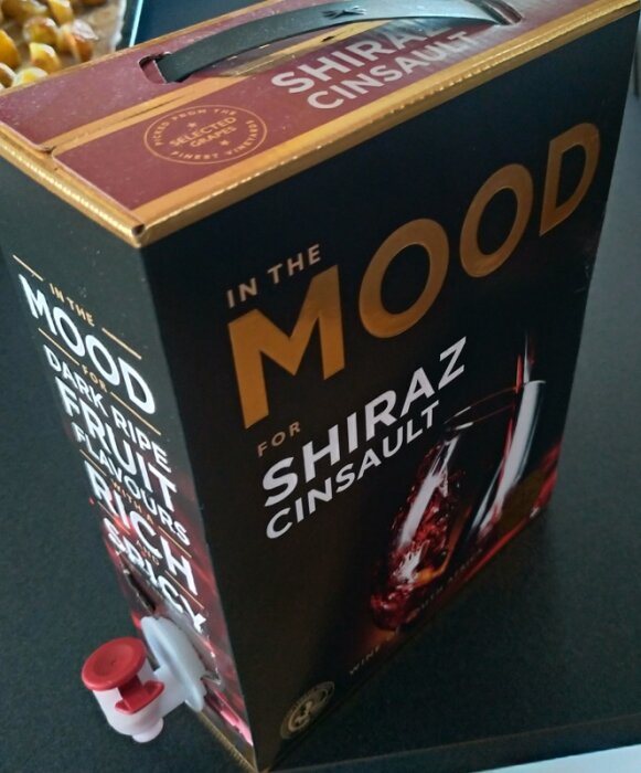 Box med Shiraz Cinsault-vin, kran, "In The MOOD," mörka fruktiga smaker, bord, bakgrund med mat.